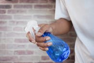 A close-up shot of a hand holding a blue spray bottle against a brick wall background.